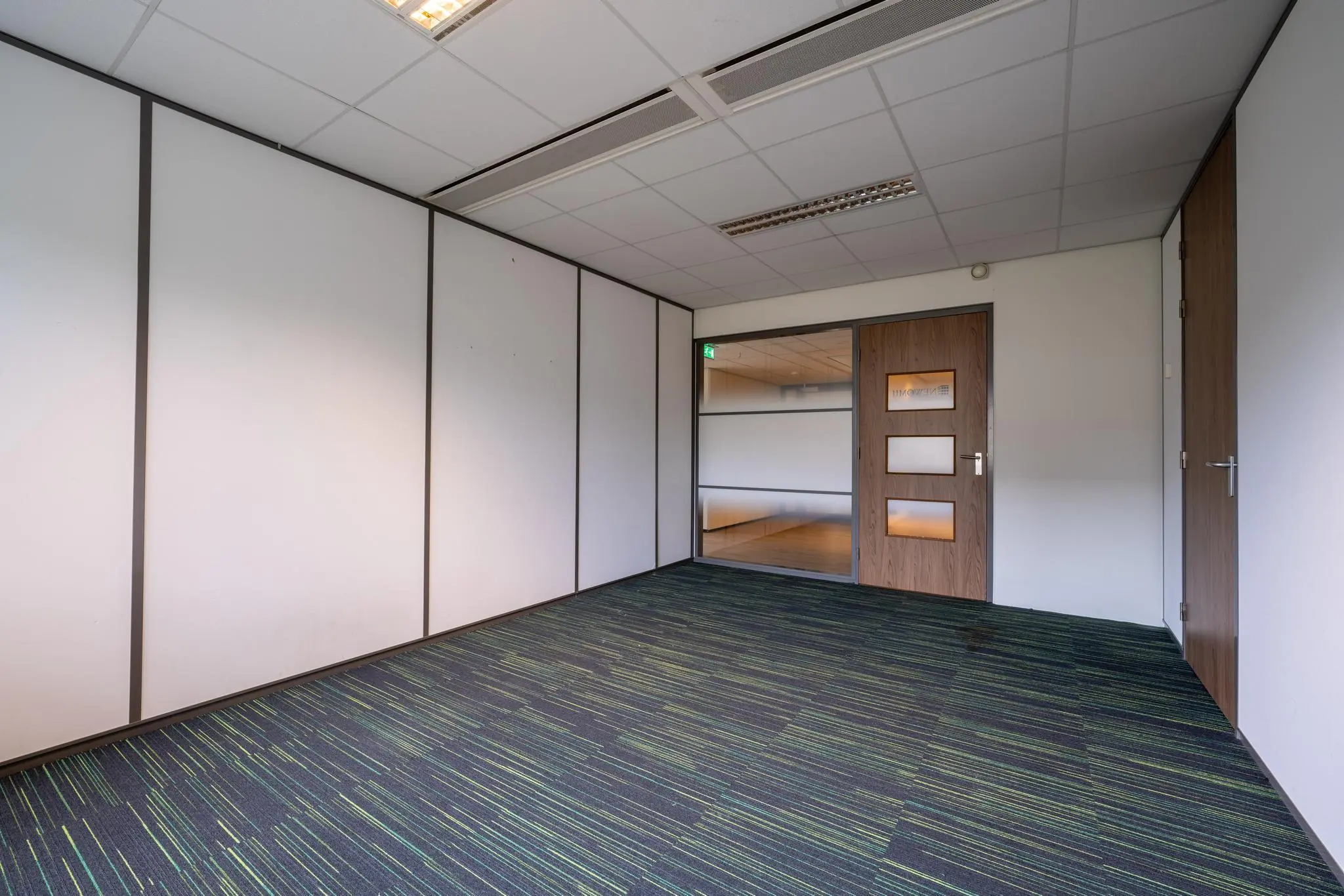 Lege kantoorruimte met witgelamelleerde wanden, een deur met glaspanelen en een vloerbedekking met blauw-groene strepen aan de Burgemeester Verderlaan.